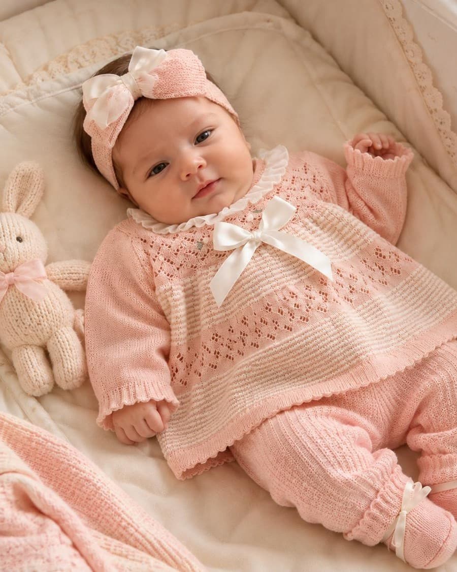 Bebé recostada en su cuna con conjunto tejido rosado, diadema con lazo y conejito de peluche tejido